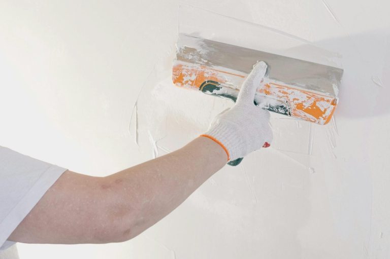 Eine Hand mit Spachtel verputzt eine weiße Wand.